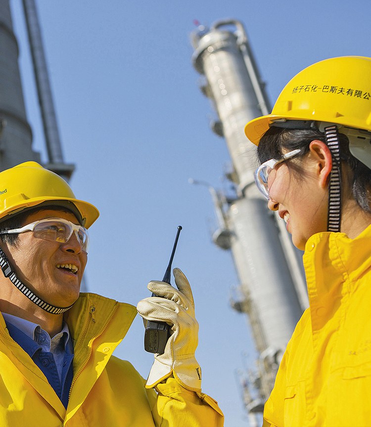 Hai người vận hành của BASF-YPC mặc áo và mũ bảo hộ màu vàng, đeo kính, đứng trước một tháp chưng cất dưới trời xanh