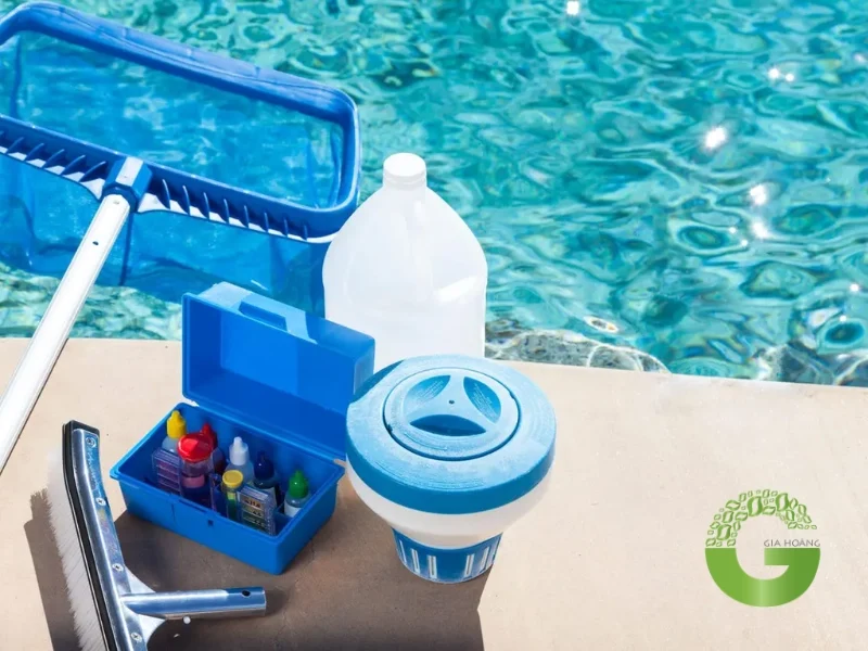 Hình ảnh chụp các dụng cụ và hóa chất được sử dụng để xử lý nước hồ bơi, bao gồm vợt vớt rác, hóa chất chlorine dạng viên, và bộ dụng cụ kiểm tra nồng độ hóa chất trong nước. Hình ảnh này minh họa quy trình bảo dưỡng và xử lý nước hồ bơi để đảm bảo nước luôn trong sạch và an toàn cho người sử dụng